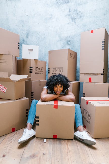 A smiling woman with curly black hair, dressed in casual clothing, is sitting on the wooden floor of an indoor space surrounded by numerous cardboard boxes of various sizes, some with printed labels and red packing tape. She is leaning on a large box placed in front of her, with her arms resting on it, and her legs extended outward, slightly apart. The boxes are stacked against the plain, textured wall, and some are partially open, revealing packing materials inside. The scene depicts a home relocation or moving process managed by Man with Van Bexleyheath, with the woman appearing to be in a staging area for furniture transport or packing and moving activities, in a well-lit room with no additional furniture visible.