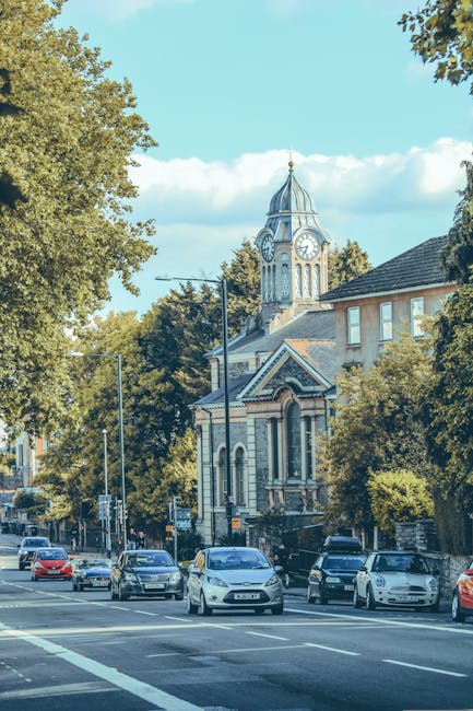 A busy street scene in Bexleyheath with several parked and moving cars along the curbside, adjacent to a sidewalk lined with mature green trees. In the background, a historic Victorian-style building with a prominent clock tower featuring a pointed roof and ornate architectural details is visible, partially obscured by lush foliage. The scene appears to be daytime with clear weather, and the area is part of a typical urban residential and commercial neighbourhood. The image reflects the environment where home relocation and furniture transport services by Man with Van Bexleyheath might operate, especially around permit and suspension zones for removals. The street is well-lit, and the overall setting conveys a typical urban environment suitable for house removals and moving logistics.