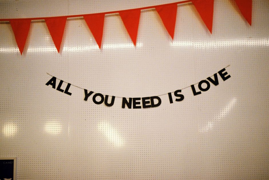 A wall decorated with a string of red triangular fabric banners hanging along the top edge and a black paper banner with white lettering reading 'ALL YOU NEED IS LOVE' suspended beneath them. The banners are in an indoor setting with soft lighting, and part of a television or monitor is visible in the lower left corner. The decorations suggest a celebratory or festive atmosphere, which could be part of a housewarming or home relocation event. This scene depicts a neatly arranged interior environment, capturing the decorative elements relevant to moving or packing celebrations. Man with Van Bexleyheath, a professional removals company, might utilize such decorative displays during packing and moving services to create a welcoming atmosphere or to mark special occasions during a home relocation in Bexleyheath.