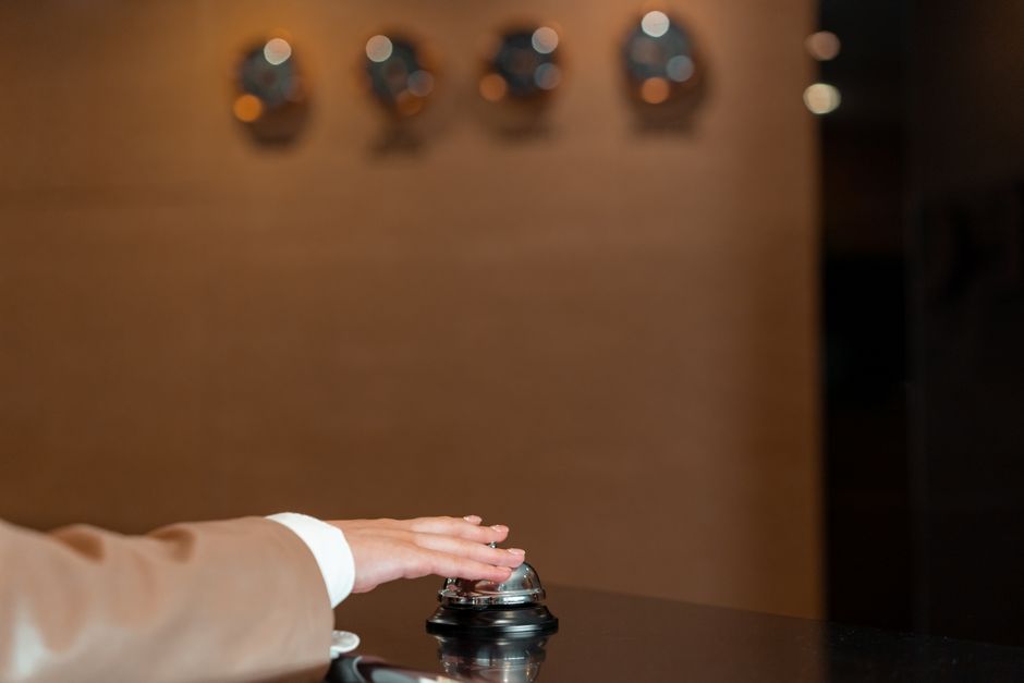A close-up of a person's hand wearing a beige suit sleeve, pressing a bell on a dark, glossy surface, likely in a house or apartment lobby, with a wooden panel and four metallic control dials visible in the blurred background. The scene suggests a key moment in home relocation processes, such as requesting access or signalling arrival during furniture transport or packing and moving activities, with the environment illuminated by warm lighting. This image may be used in the context of house removals or relocation services provided by Man with Van Bexleyheath, illustrating aspects of the logistics involved in loading or entering a property for a professional removal or moving service.