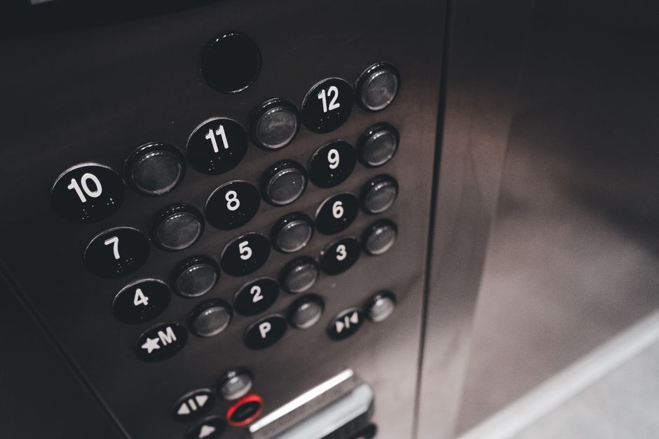 A person wearing a grey sweatshirt and carrying a black backpack is pressing a button on the interior control panel of an elevator, which is situated inside a residential building. The elevator's doorframe is metallic, and the control panel features numbered buttons, including the ground floor and other levels, as well as an emergency or service button. The scene is illuminated with neutral lighting, emphasizing the context of home relocation or furniture transport. The individual appears to be preparing to select a floor for moving large boxes, furniture, or appliances as part of a house removal process, supporting the services offered by Man with Van Bexleyheath, located inside the property near the doorway or elevator entrance.