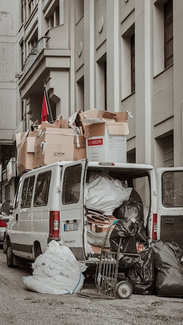 A white commercial van parked on a city street with its rear doors open, revealing various stacked cardboard boxes, some open and others sealed with tape, along with large plastic bags filled with packing materials and miscellaneous items. The boxes are of different sizes, with some marked for disposal. A small hand trolley and a few black rubbish bags are positioned near the open van, indicating a loading or unloading process. The van is situated beside a multi-storey building with large windows and light-colored stone or concrete facade, typical of urban residential or commercial properties. The scene captures part of the street environment, including pavement and adjacent building structures. This image illustrates a home relocation or clearance activity related to professional removals services, such as those provided by Man with Van Bexleyheath, highlighting the loading process for a house move or bulky waste removal during a move in Bexleyheath.