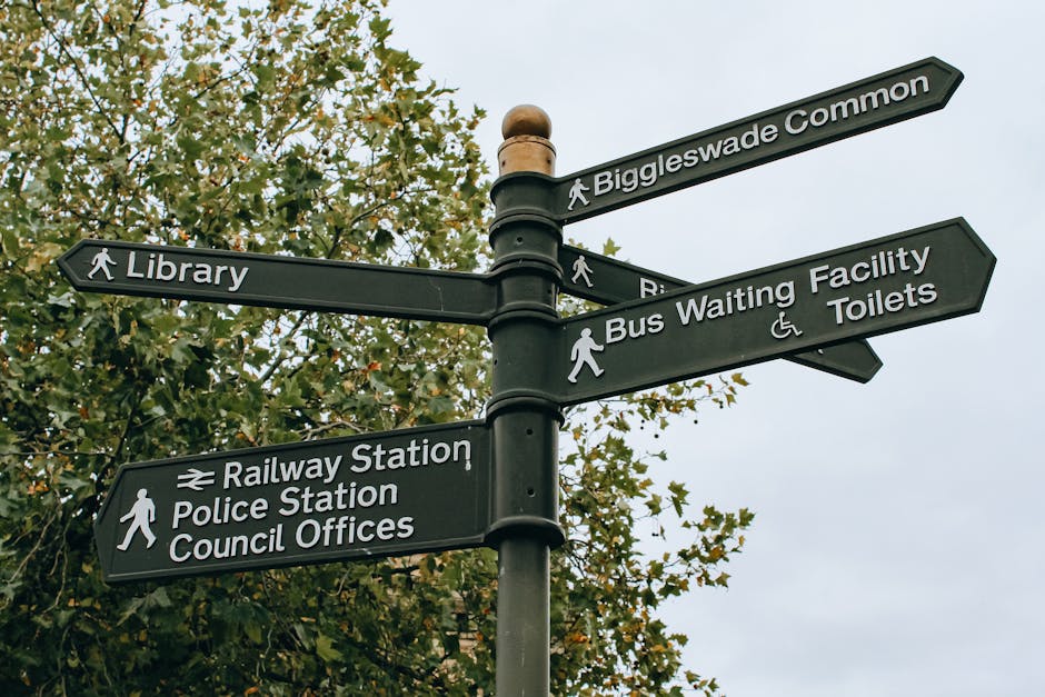 A black metal signpost with multiple directional signs attached, situated outdoors against a backdrop of green leafy trees and an overcast sky. The signs display white text indicating directions to local amenities, including 'Biggleswade Common', 'Library', 'Railway Station', 'Police Station', 'Council Offices', 'Bus Waiting Facility', and 'Toilets.' The signpost is positioned on a paved pavement, with the signs extending horizontally in different directions, guiding pedestrians and visitors around the area. In the context of home relocation and moving logistics, such directional signage can be relevant for planning collection or delivery points when coordinating furniture transport or packing and moving services offered by [COMPANY_NAME], especially in areas with narrow streets or complex access routes that require careful navigation during a house removal process.