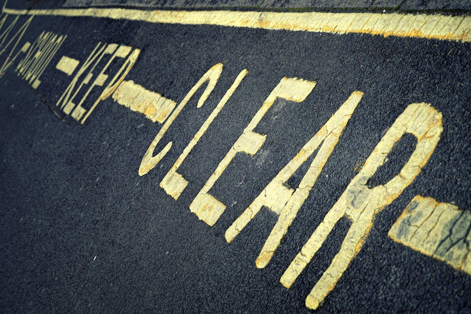Close-up of a weathered road surface showing yellow painted words 'CLEARWAY' on dark asphalt, with some paint chipping and cracking visible. The image includes a portion of a yellow line above the text, indicating a no-parking zone typically found in street parking restrictions. The background features an out-of-focus curb and part of a residential street, suggesting the scene is in a neighbourhood where vehicle loading and unloading are regulated. This scene could relate to parking and permit advice for house removals in the context of home relocation services by Man with Van Bexleyheath, highlighting the importance of understanding street and parking regulations during furniture transport and moving logistics in residential areas such as Bexleyheath.
