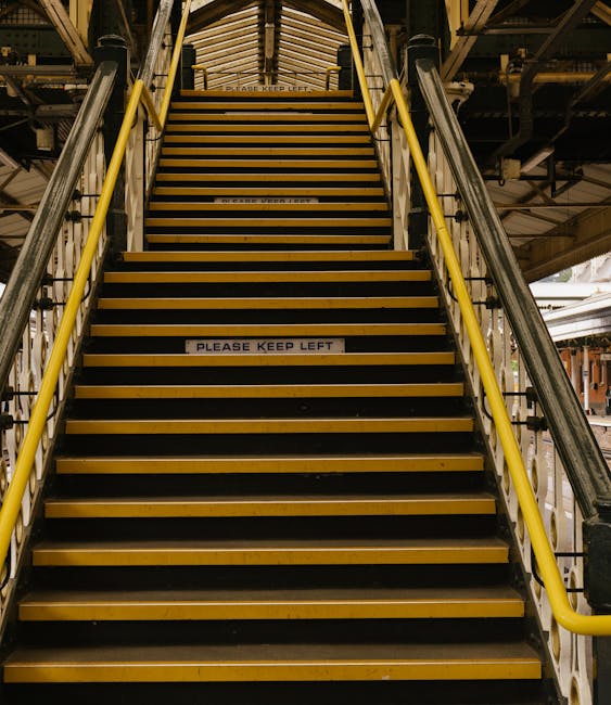 A downward view of a concrete staircase inside a building leading to a dark basement or lower level. The staircase is enclosed by beige tiled walls with a blue horizontal stripe near the top. An orange metal handrail runs along both sides of the stairs, and the steps are made of concrete with metal non-slip strips. Sunlight casts shadows on the stairway, partially illuminating the upper steps and the tiled wall. The image is taken from the top of the stairs, looking down towards the basement or lower level entrance, which appears to be an open, dark doorway. This setting is typical of communal or public building staircases involved in house removals or moving logistics, such as loading or unloading areas. The photograph aligns with house removals and furniture transport activities, emphasizing the environment where careful handling and navigation are essential for successful home relocation processes, as provided by Man with Van Bexleyheath.