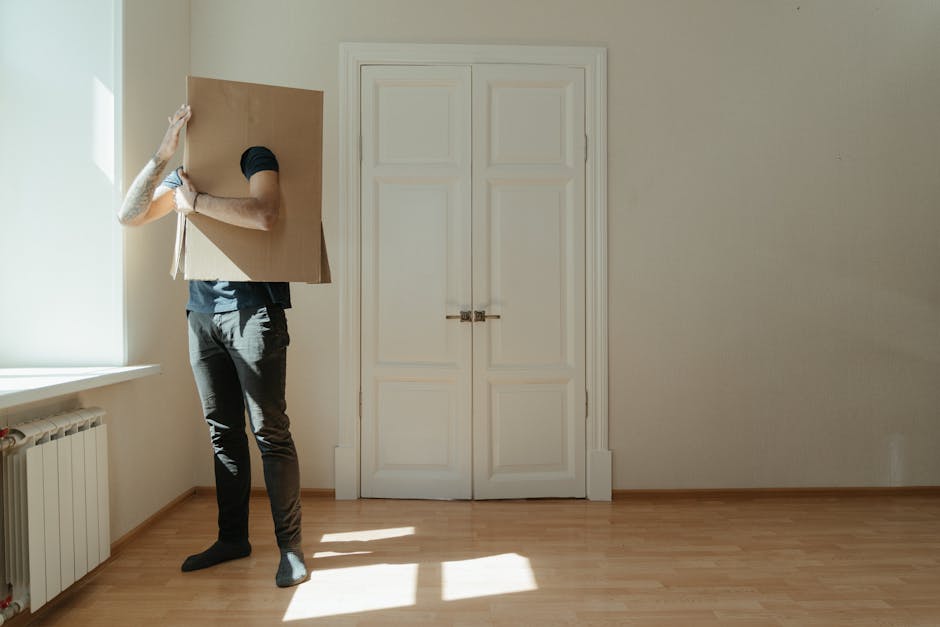 A person dressed in dark clothing and wearing black gloves is carrying a large, flat cardboard box in a well-lit room with light-colored wooden flooring. The individual is holding the box securely with both hands, with their right arm visible and tattoos on their forearm. The room features a white double door with a silver latch in the background, closed, and a white radiator located beneath a window on the left side, allowing natural light to illuminate the space. The window frame is white, and the walls are plain and light-colored. The scene captures part of a home relocation process, with the person preparing to move the box through a typical residential interior, supported by [COMPANY_NAME] during the packing and moving stages as part of their removals service for house or flat moves, especially in tight hallways or narrow spaces.