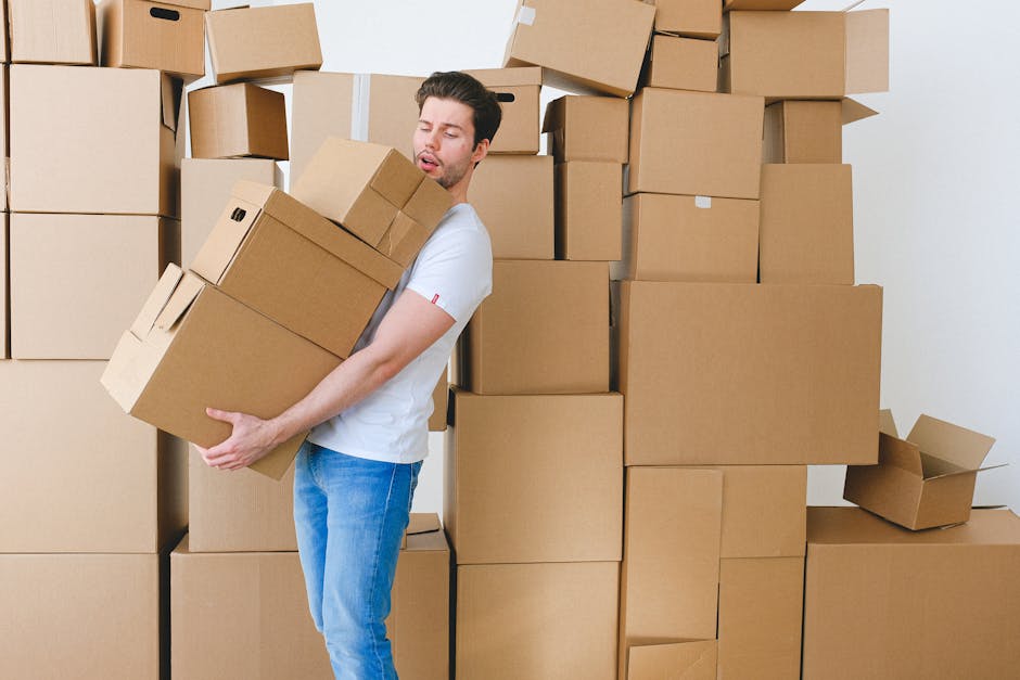 A man in casual clothing, including a white t-shirt and blue jeans, is inside a room filled with numerous cardboard boxes of various sizes, some sealed with packing tape and others open, revealing packing materials such as tissue paper and plastic wrap. He appears to be lifting or carrying a medium-sized box, with his eyes closed and an expression of effort or concentration. The background shows a wall with more stacked boxes, indicating an active home relocation or furniture transport process. The scene is well-lit with natural or interior lighting, emphasizing the organized packing as part of the packing and moving phase. The image includes the environment of a typical room used for packing during house removals, aligning with services offered by [COMPANY_NAME], such as removals and transportation in Bexleyheath.