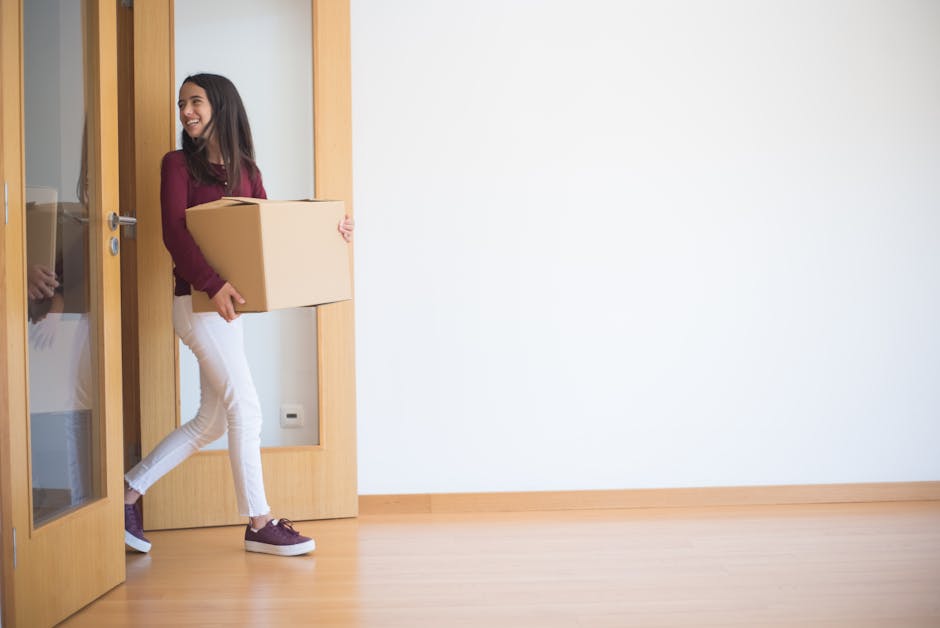 A person dressed in dark clothing and wearing black gloves is carrying a large, flat cardboard box in a well-lit room with light-colored wooden flooring. The individual is holding the box securely with both hands, with their right arm visible and tattoos on their forearm. The room features a white double door with a silver latch in the background, closed, and a white radiator located beneath a window on the left side, allowing natural light to illuminate the space. The window frame is white, and the walls are plain and light-colored. The scene captures part of a home relocation process, with the person preparing to move the box through a typical residential interior, supported by [COMPANY_NAME] during the packing and moving stages as part of their removals service for house or flat moves, especially in tight hallways or narrow spaces.