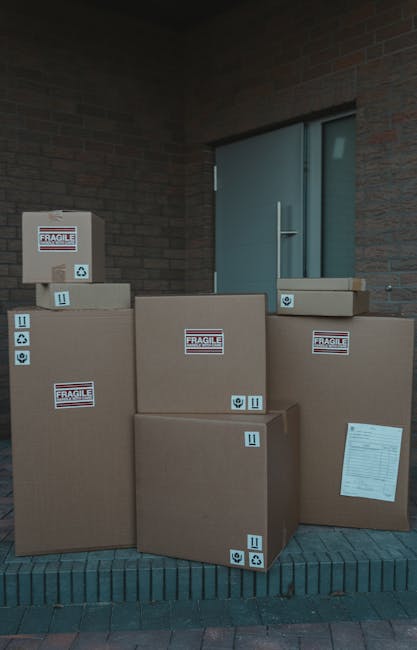 A collection of six large cardboard boxes, some labeled with 'FRAGILE' stickers and handling symbols, are stacked outside a modern brick building near a blue door with a metal handle. The boxes vary in size, with some placed directly on a paved surface and others leaning against the building wall. One box has a printed label visible on the front. The background shows a brick wall and a slightly open door, indicating a residential or commercial property. The scene suggests preparations for a home or office move, with the boxes ready for transport as part of a furniture transport or packing and moving process undertaken by a professional removal service such as Man with Van Bexleyheath.