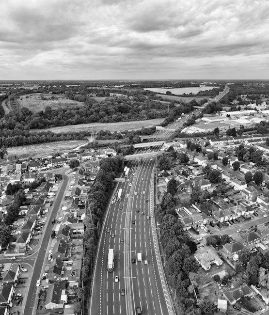 An aerial black-and-white photograph displays a suburban residential area adjacent to a major multi-lane highway. The scene includes rows of houses with small gardens, driveways, and trees lining the streets. The highway features moving vehicles such as cars and trucks, with visible lane markings and a central divider. Beyond the urban area, open fields, patches of woodland, and a river or large water body can be seen extending toward the horizon. The sky is filled with scattered clouds, and the overall setting suggests daytime. In the context of house removals and relocation services like those offered by Man with Van Bexleyheath, the image subtly reflects the logistics of home relocation and the importance of efficient transport infrastructure in moving services.