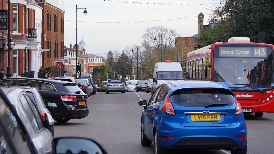 A busy urban street scene with multiple parked and moving vehicles, including a black car, a silver car, and a white van positioned on the street. In the foreground, a blue hatchback with a UK registration plate is parked close to the curb, adjacent to a red bus displaying route information for 'Brent Cross 143' and operated by Metroline. Behind the vehicles, residents and pedestrians are walking along the sidewalk lined with traditional red-brick and white buildings, some with decorative architectural features. Street lamps and overhead electric wires extend across the scene, while trees with beginning to show autumn foliage are visible in the background. The weather appears overcast with diffuse natural light illuminating the scene, which reflects typical city planning and moving logistics relevant to home relocation and furniture transport services offered by Man with Van Bexleyheath. This image exemplifies the typical environment faced during removals and packing and moving operations in the Bexleyheath area, highlighting street conditions and vehicle positioning involved in residential relocations.
