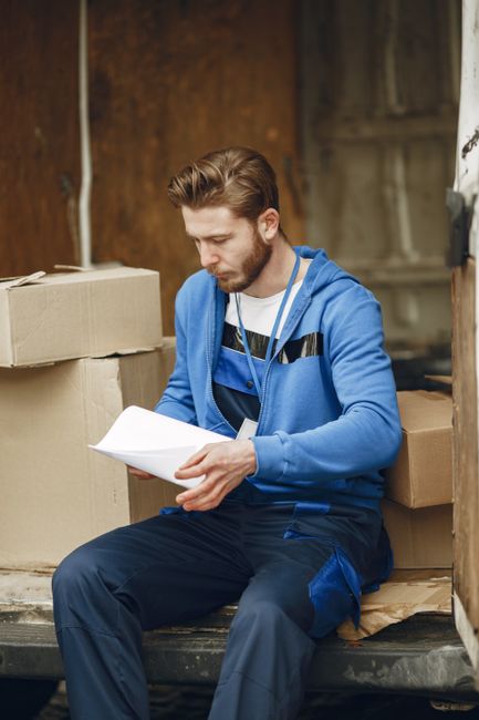 A young man with styled brown hair and a beard is sitting on the edge of a moving truck or storage area, surrounded by stacked cardboard boxes of varying sizes, some taped and others open. He is weari