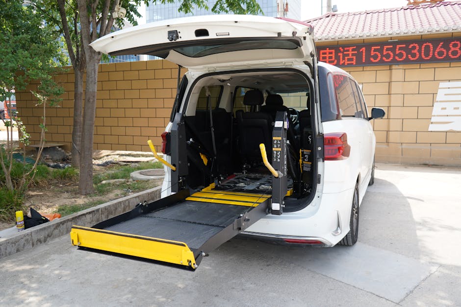 A white vehicle with its rear hatch open, revealing a wheelchair lift platform extended onto the pavement for a home relocation or furniture transport process. Inside the vehicle, visible are black seats and safety straps, with the wheelchair lift featuring yellow safety rails and a textured surface for secure loading. The surrounding environment includes a brick wall with a red roof, a sign with red text, a tree providing some shade, and a concrete curb along the edge of the parking area. The vehicle is parked on an asphalt surface near a building, and the scene is lit by natural daylight, indicating a clear and bright day. This setup demonstrates professional packing and loading procedures typically carried out by a removals service, such as Man with Van Bexleyheath, supporting efficient and accessible home moving operations.